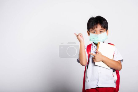 Téléchargez les photos : Asiatique étudiant enfant garçon porter étudiant thai uniforme et protéger masque prêt à aller à l'école pointant du doigt vers l'espace en studio tourné isolé sur fond blanc, préscolaire, nouveau normal de retour à l'école - en image libre de droit