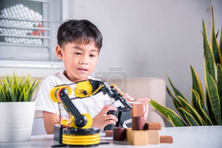Téléchargez les photos : Heureux petit garçon asiatique utilisant la télécommande jouant bras de machine robotique pour ramasser le bloc de bois, drôle enfant apprenant avec succès obtenir leçon bras robot de contrôle, éducation de la science de la technologie - en image libre de droit