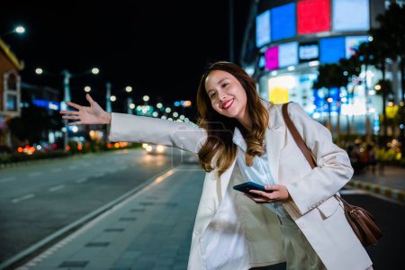 Foto de Asian businesswoman standing hail waving hand taxi on road in busy city street at night, beautiful woman smiling using mobile phone application hailing with hand up calling cab after late work - Imagen libre de derechos
