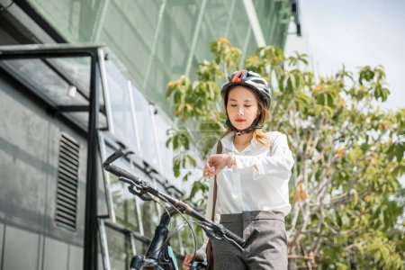 Foto de Retrato sonriente mujer reloj de verificación, joven mujer de negocios asiática de pie con bicicleta comprobar el tiempo en los edificios modernos calle de la ciudad por la mañana antes de ir a trabajar en la oficina, viajero de negocios, estilo de vida - Imagen libre de derechos