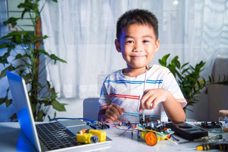 Foto de El niño pequeño aprende remotamente en línea con el juguete del coche antes de enviar el código, el niño asiático enchufa la energía y el cable de la señal al microprocesador del sensor con el coche del robot de Arduino, aprendizaje de la tecnología de la IA de la educación de STEAM - Imagen libre de derechos