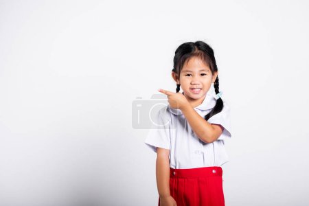 Photo for Asian toddler smiling happy wear student thai uniform red skirt keeps pointing finger at copy space in studio shot isolated on white background, Portrait little children girl preschool, Back to school - Royalty Free Image