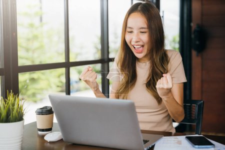 Photo for Excited happy Asian business young woman sitting with laptop computer at cafe desk raising hands up and celebrating success, female feeling winner rejoicing online win got new job at home - Royalty Free Image