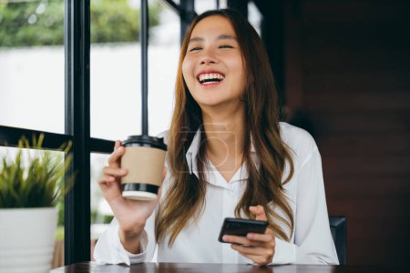 Foto de Asiática joven mujer de negocios sosteniendo taza de papel de café y el uso de teléfono móvil para chating en línea en la cafetería, Mujer feliz sentado en la cafetería cerca de la ventana que sostiene el teléfono móvil en la mano - Imagen libre de derechos