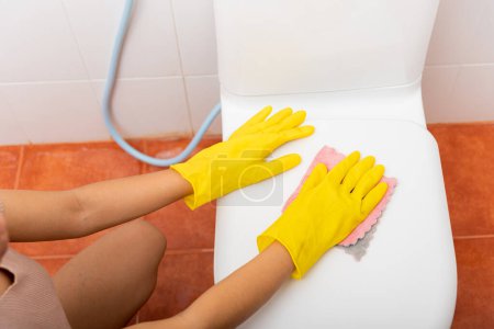 Foto de Las manos de la mujer asiática limpiando el asiento del inodoro con un paño rosa limpian el baño en la casa, la mujer usa guantes de goma amarillos, se sienta y limpia o lava el baño, concepto de atención médica del ama de llaves - Imagen libre de derechos