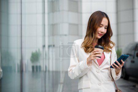Téléchargez les photos : Femme d'affaires asiatique tenant tasse de café à emporter regarder smartphone va travailler marcher sur la rue de la ville près de bureau le matin, Portrait femme d'affaires tenir téléphone mobile et tasse en papier de boisson chaude en plein air - en image libre de droit