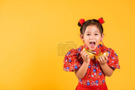 Photo for Chinese New Year. Happy Asian Chinese little girl smile wearing red cheongsam holding gold ingot, Portrait children in traditional dress hold golden bar, studio short isolated on yellow background - Royalty Free Image