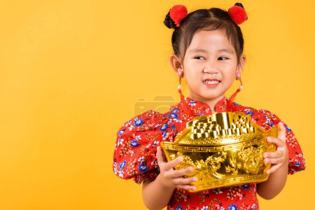 Photo for Chinese New Year. Happy Asian Chinese little girl smile wearing red cheongsam holding big gold ingot, Portrait children in traditional dress hold golden bar, studio short isolated on yellow background - Royalty Free Image