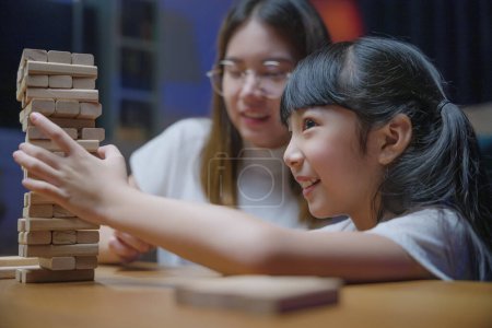 Téléchargez les photos : Asiatique jeune mère jouer jeu en bloc de bois avec sa petite fille dans le salon à la maison la nuit, femme souriante aider à enseigner jeu d'enfant construire constructeur de blocs de bois, éducation - en image libre de droit
