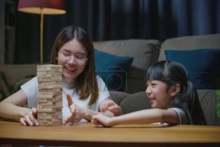 Photo for Smiling woman help teach child play build constructor of wooden blocks, Asian young mother playing game in wood block with her little daughter in home living room at night time before going to bed - Royalty Free Image