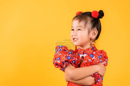 Téléchargez les photos : Happy Asian Chinese little girl smile wear red cheongsam crossarms, Portrait enfants en robe traditionnelle avec célébration de bienvenue pour le Nouvel An chinois, studio court isolé sur fond jaune - en image libre de droit
