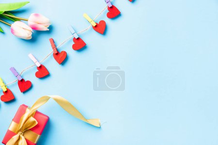 Téléchargez les photos : Joyeux concept de Saint Valentin. décoration de valentines en forme de coeur rouge accrochée avec des clips pour l'amour et boîte cadeau rouge avec ruban d'or sur la carte de vœux de corde sur fond bleu avec espace de copie - en image libre de droit