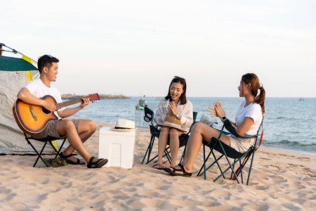 Photo for Happy friend have fun playing guitar and clap in camp they smiling together in holiday on sand beach near camping tent vacation time at sunset, Young Asian group woman and man in summer travel outdoor - Royalty Free Image