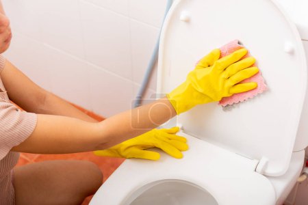 Téléchargez les photos : Mains de femme asiatique nettoyage siège de toilette par chiffon rose essuyer les toilettes à la maison, femme portant des gants en caoutchouc jaune, elle est assise et nettoyer ou laver la salle de bain, concept de soins de santé de femme de ménage - en image libre de droit