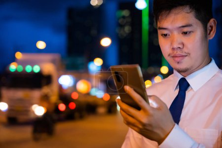 Photo for Young man walking and chatting on mobile phone with friends at social networks outdoor, Portrait Asian businessman typing an sms message via smartphone after work near office at night city street - Royalty Free Image