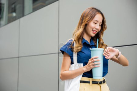 Foto de Asian beautiful woman confident smiling with cloth bag holding steel thermos tumbler mug water glass she walking outdoors on street near modern building office, Happy female looking watch time - Imagen libre de derechos