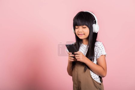 Foto de Niño asiático 10 años disfrutando de escuchar música desde el teléfono móvil usar auriculares inalámbricos ojos cerrados en el estudio tiro aislado sobre fondo rosa, niña feliz uso de teléfono inteligente escuchar música con auriculares - Imagen libre de derechos