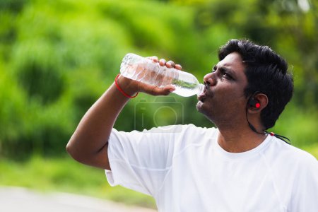 Foto de Primer plano asiático joven corredor deportivo negro hombre usar auriculares atleta que beber agua de una botella después de correr en el parque de salud de la calle al aire libre, concepto de ejercicio saludable - Imagen libre de derechos
