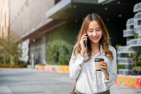 Téléchargez les photos : Asiatique jeune femme d'affaires marchant à l'extérieur parlant sur téléphone portable intelligent et tenant tasse de café à emporter, mode de vie femme d'affaires tenir tasse en papier et appelant smartphone à l'extérieur du bureau - en image libre de droit