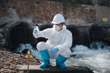 Ecologista toma de muestras de productos químicos tóxicos del agua del río con vidrio de tubo de ensayo y tienen humo blanco, Biólogo usar traje de protección y máscara recoge muestras de aguas residuales de industrial, ambiente problemático
