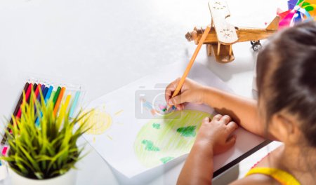 Téléchargez les photos : Asiatique mignon enfant préscolaire assis sur la table souriant elle dessine image avec crayon à la maison, Happy child petite fille dessin coloré famille debout tenir la main sur la planète terre sur papier, concept de jour de la terre - en image libre de droit