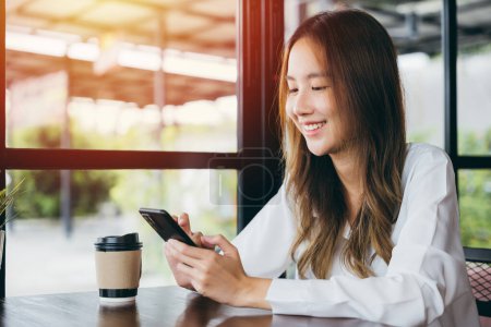 Photo for Happy female lifestyle sitting in cafe near window she cellphone on mobile phone, Asian young business woman freelance holding smartphone and coffee paper cup at coffee shop - Royalty Free Image