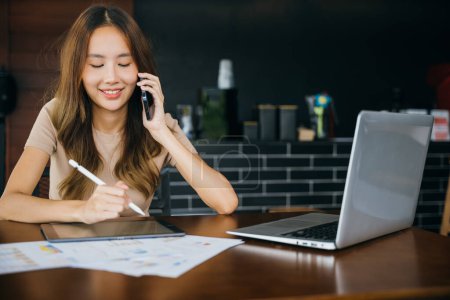 Foto de Mujer de negocios asiática independiente sentada en la cafetería llamada teléfono móvil hablando y preguntando sobre algo, sonriendo computadora portátil de trabajo femenino y tomar nota en la tableta digital en la oficina en casa - Imagen libre de derechos