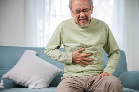 Älterer asiatischer alter Mann sitzt mit Bauchschmerzen auf dem Sofa, hohes Alter leidet unter Bauchschmerzen, hält seinen Bauch im Wohnzimmer, Menschen Gesundheitsproblem, Lebensmittelvergiftung