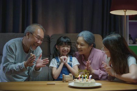 Foto de Feliz familia asiática abuelos y madre celebrando disfrutar cantando cumpleaños canción fiesta con pastel de nieta soplando vela en casa en la noche, cumpleaños sorpresa concepto - Imagen libre de derechos