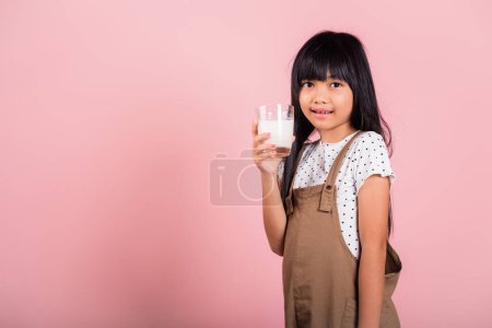 Téléchargez les photos : Asiatique petit enfant 10 ans sourire tenir lait verre boisson lait blanc au studio tourné isolé sur fond rose, Happy child girl vie quotidienne soins de santé nourriture - en image libre de droit