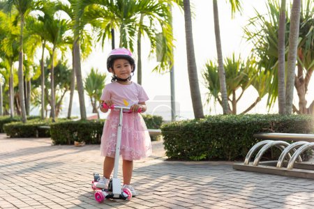 Téléchargez les photos : Scooter enfant. Joyeux Asiatique petite fille enfant porter un casque sûr en jouant rose kick board sur la route dans le parc à l'extérieur le jour de l'été, Jeux actifs pour enfants à l'extérieur, Sport pour enfants concept de mode de vie sain - en image libre de droit