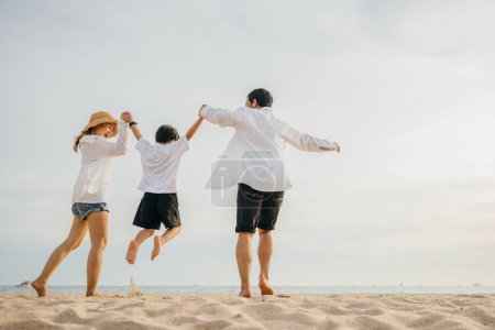 Téléchargez les photos : Heureuse famille asiatique avoir du plaisir et de vivre un mode de vie sain ensemble sur la plage, Dos famille mère, père et fils se tenant la main et sautant dans l'air à l'aube, concept d'activités de plein air de la famille - en image libre de droit