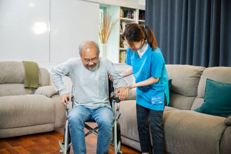 Photo for Doctor support old man to getting up to exercise, help handicapped elderly stand up, Asian woman nurse helping senior man patient get up from wheelchair for practice walking at home, physical therapy - Royalty Free Image