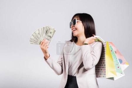 Photo pour Femme avec des lunettes sourire client confiant tenant sacs à provisions en ligne multicolore et billets d'argent en dollars sur la main, heureux heureux asiatique jeune femme studio tourné isolé sur fond blanc - image libre de droit