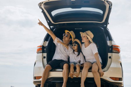 Photo for Tourism Day. Dad, mom and daughter enjoying road trip sitting on back car and pointing finger out blue sky, people having fun in summer vacation on beach, Family traveling in holiday at sea beach - Royalty Free Image