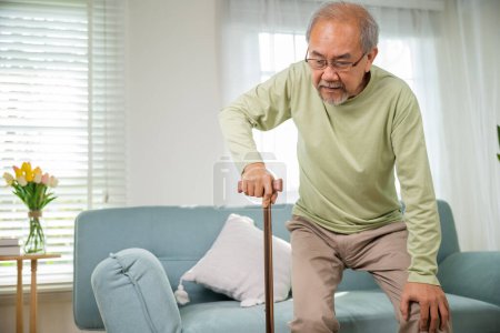 Photo for Asian Old man with eyeglasses typing to stand up from sofa with walking cane, Elderly suffering from knee pain ache holding handle of cane, senior disabled man holding walking stick at home - Royalty Free Image