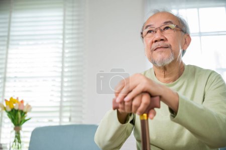 Foto de Las manos de un anciano asiático sentado descansando en casa sostienen un bastón de madera para caminar, la mano anciana sostiene el mango del bastón, el hombre discapacitado mayor sostiene el bastón, el concepto de atención médica de jubilación - Imagen libre de derechos