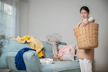 Téléchargez les photos : Belle femme de ménage faisant des travaux ménagers tient panier en bois de vêtements sales et propres à laver à la maison, femme au foyer asiatique tenant panier avec tas de vêtements différents sur le canapé dans le salon - en image libre de droit