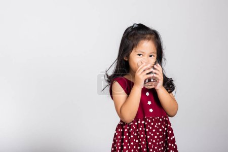 Téléchargez les photos : Petite fille mignonne enfant 3-4 ans sourire boire de l'eau fraîche à partir de verre en studio tourné isolé sur fond blanc, Asiatique enfants d'âge préscolaire, Vie quotidienne santé - en image libre de droit