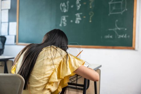 Photo for Education. Back view of school girl on lesson in classroom write hardworking on blackboard, primary child is sitting lessons at table in school writing or drawing in notebook, Back to school concept - Royalty Free Image