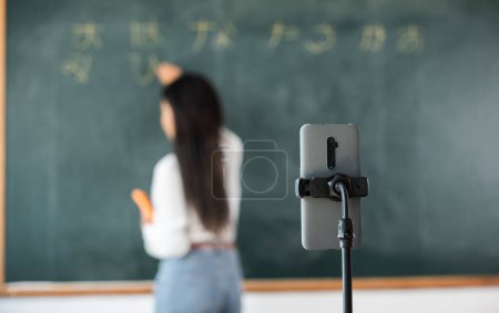 Photo for Back to school online class. Asian teacher teach on board while have blackboard lecture online through smartphone in classroom, woman standing front of class live video call online smart mobile phone - Royalty Free Image