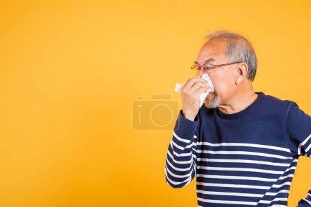 Foto de Pensionista enfermo. Asiático anciano resfriado teniendo gripe y estornudos de enfermedad virus problema uso papel tisú, retrato anciano viejo soplado nariz con tejido estudio disparo aislado en amarillo fondo - Imagen libre de derechos