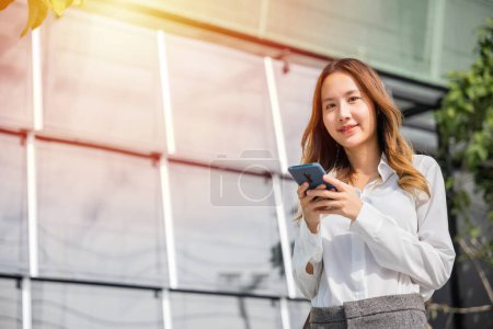Téléchargez les photos : Happy young woman using smartphone outdoor on urban city street, applications on cell phone, people checking social media, Asian businesswoman holding mobile phone texting message outside of office - en image libre de droit
