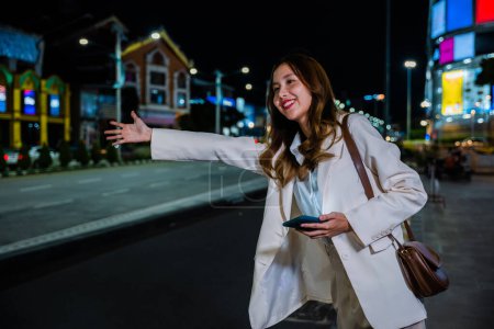 Foto de Hermosa mujer sonriendo usando aplicación de teléfono inteligente que llama con la mano hacia arriba taxi después del trabajo tardío, mujer de negocios asiática de pie granizo saludando taxi de la mano en la carretera en la calle de la ciudad ocupada por la noche - Imagen libre de derechos