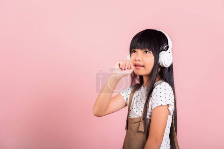 Téléchargez les photos : Asian little kid 10 years old smile listening music wear wireless headset and keeps hand near mouth sings song, studio shot isolated on pink background, Child girl funny listen music with headphones - en image libre de droit