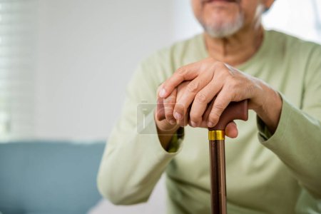 Téléchargez les photos : Mains de vieil homme asiatique assis au repos à la maison tenir canne à pied en bois, main âgée tenant poignée de canne, homme handicapé âgé tenant bâton de marche, concept de soins médicaux de retraite - en image libre de droit