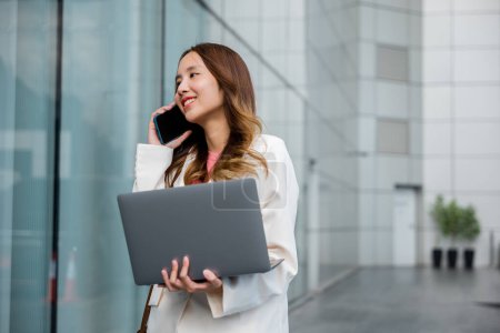 Photo for Portrait beautiful business woman smiling holding computer discussing issues on smart mobile phone in city, Asian businesswoman working on laptop and talking cell phone at front building near office - Royalty Free Image