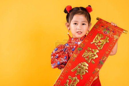 Téléchargez les photos : Chinese New Year. Happy Asian Chinese little child girl smile wearing red cheongsam qipao Spring festival couplets Character FU means fortune, blessing, studio short isolated on yellow background - en image libre de droit