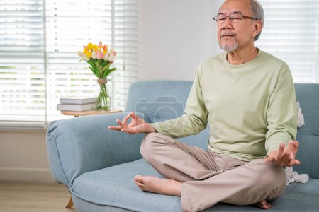 Téléchargez les photos : Vieil homme asiatique pratiquer le yoga et la méditation en position de lotus et les yeux fermés, style de vie senior pose de lotus faire du yoga pour l'équilibre mental respiration air relaxant sur le canapé à la maison, habitudes de vie saines - en image libre de droit