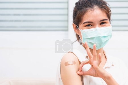 Foto de Asian young woman smile shes have adhesive plaster on arm her vaccinated and showing OK finger sign after getting immunity vaccine COVID-19 prevent - Imagen libre de derechos